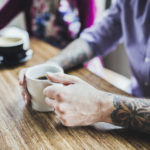 A trendy hipster man and woman with tattoos smile and talk as they enjoy a coffee and latte at a bright cafe, sitting at a large bamboo wood table.  Vertical overhead shot with copy space.